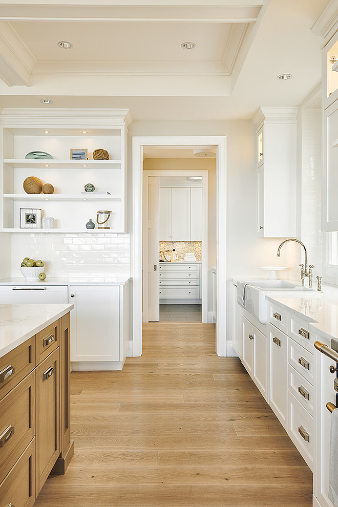This is an example of a large beach style l-shaped eat-in kitchen in Vancouver with a farmhouse sink, shaker cabinets, white cabinets, granite benchtops, white splashback, ceramic splashback, coloured appliances, light hardwood floors, with island, beige floor and white benchtop.