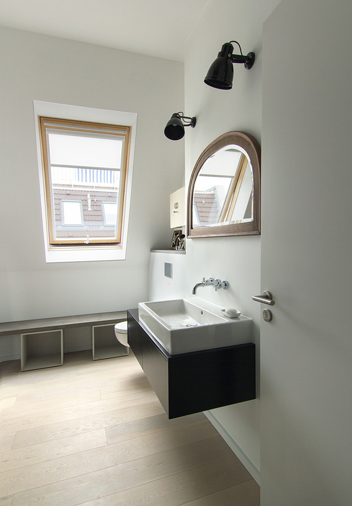 Mid-sized industrial 3/4 bathroom in Berlin with flat-panel cabinets, black cabinets, white walls, medium hardwood floors, a vessel sink, a curbless shower, a wall-mount toilet, black tile, porcelain tile, solid surface benchtops, beige floor and an open shower.