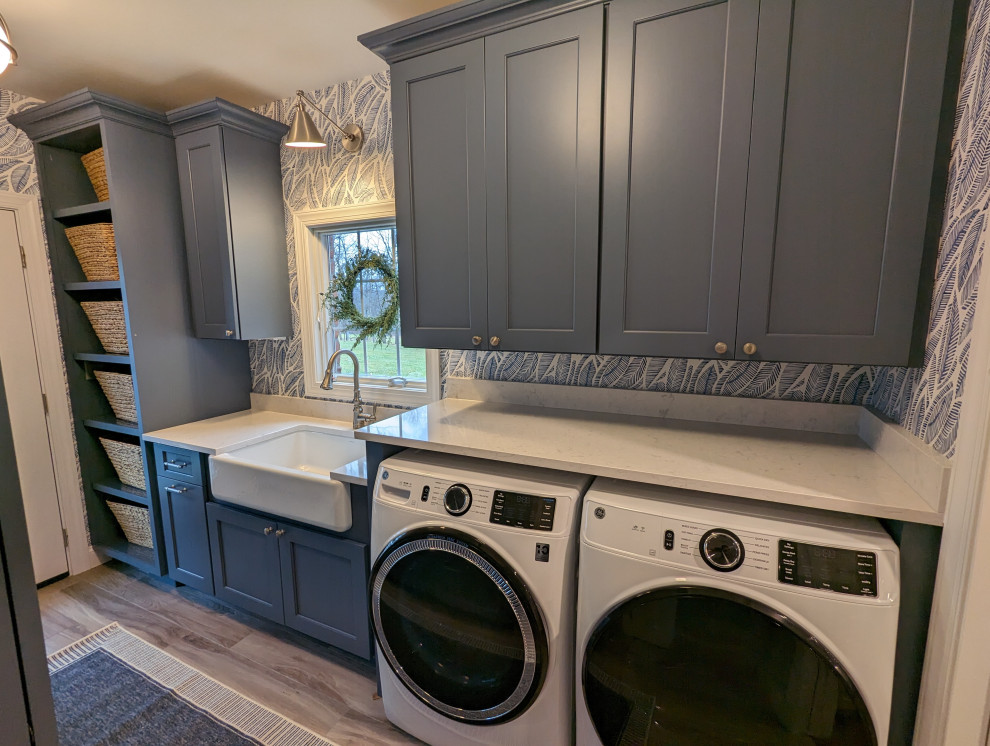 Mid-sized trendy galley gray floor and wallpaper dedicated laundry room photo in Baltimore with a farmhouse sink, recessed-panel cabinets, blue cabinets, quartz countertops, blue walls, a side-by-side washer/dryer and white countertops