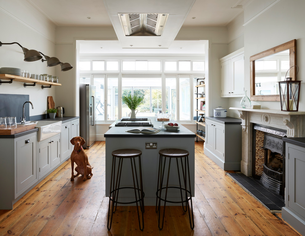 Mid-sized transitional open plan kitchen in Essex with a farmhouse sink, shaker cabinets, grey cabinets, black splashback, medium hardwood floors, with island, brown floor and black benchtop.