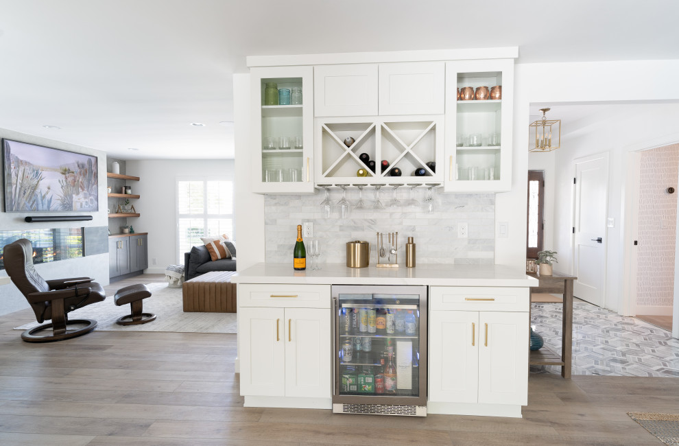 Photo of a mid-sized transitional galley home bar in Orange County with no sink, shaker cabinets, white cabinets, quartz benchtops, grey splashback, stone tile splashback, vinyl floors, brown floor and white benchtop.