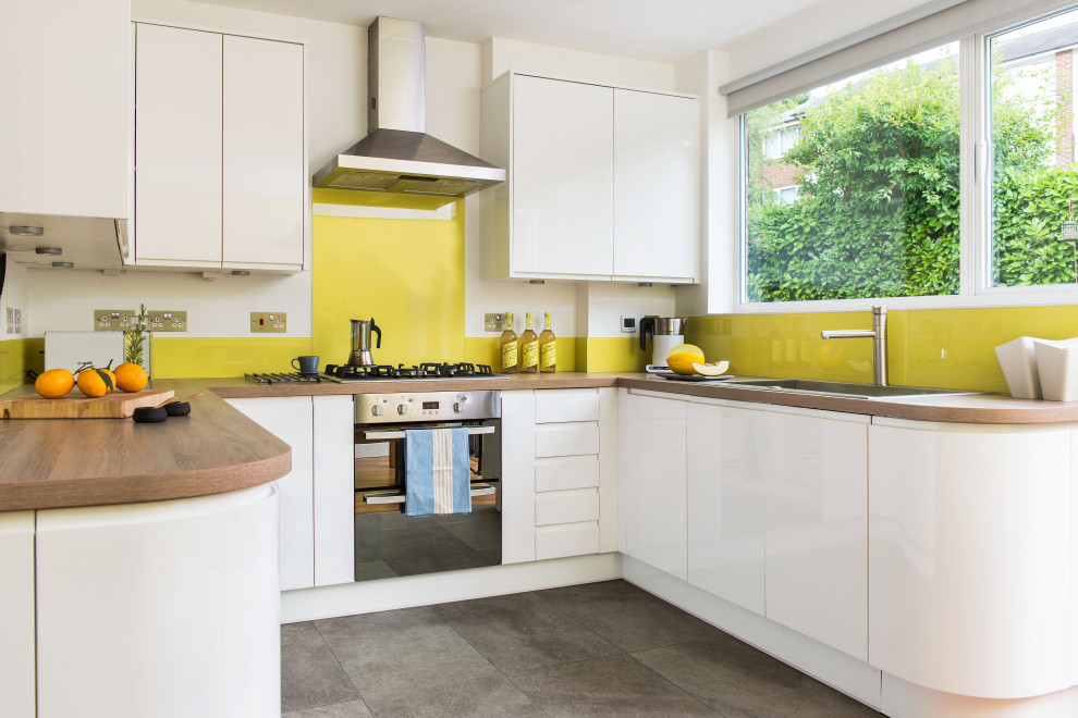Design ideas for a medium sized contemporary u-shaped open plan kitchen in London with flat-panel cabinets, white cabinets, wood worktops, yellow splashback, glass sheet splashback, grey floors, brown worktops, a built-in sink, stainless steel appliances and a breakfast bar.