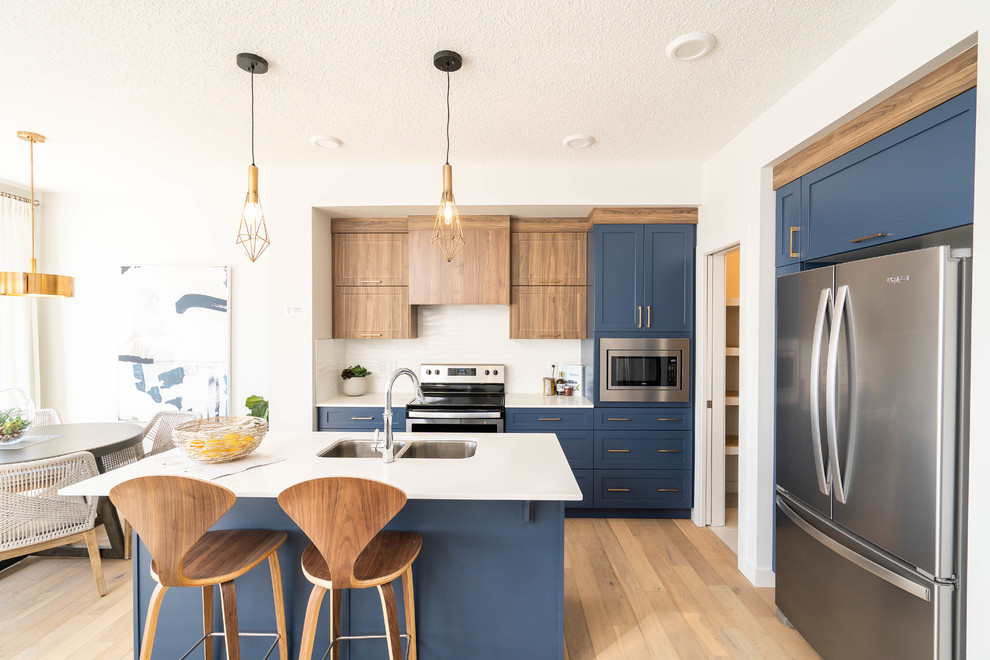 Example of a small trendy l-shaped light wood floor and beige floor open concept kitchen design in Edmonton with shaker cabinets, blue cabinets, white backsplash, stainless steel appliances, an island, white countertops, an undermount sink, marble countertops and ceramic backsplash