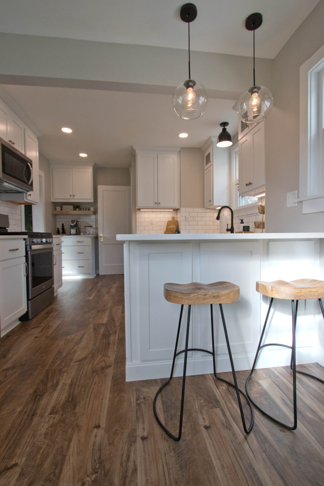 Mid-sized mountain style galley vinyl floor and brown floor enclosed kitchen photo with a farmhouse sink, recessed-panel cabinets, white cabinets, quartz countertops, white backsplash, subway tile backsplash, stainless steel appliances, a peninsula and white countertops