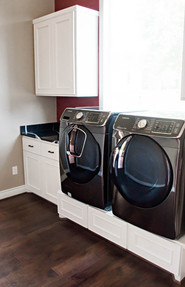 Small modern single-wall dedicated laundry room with an undermount sink, recessed-panel cabinets, white cabinets, quartz benchtops, black splashback, engineered quartz splashback, grey walls, medium hardwood floors, a side-by-side washer and dryer, brown floor and black benchtop.