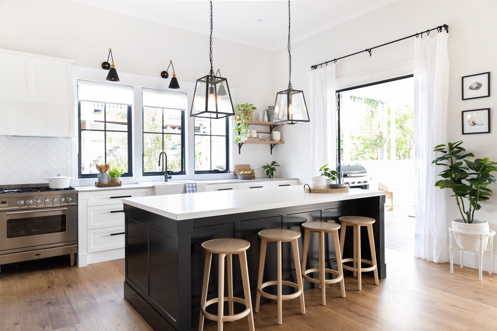 Country kitchen in Brisbane with a farmhouse sink, shaker cabinets, white splashback, stainless steel appliances, medium hardwood floors, with island, brown floor and white benchtop.