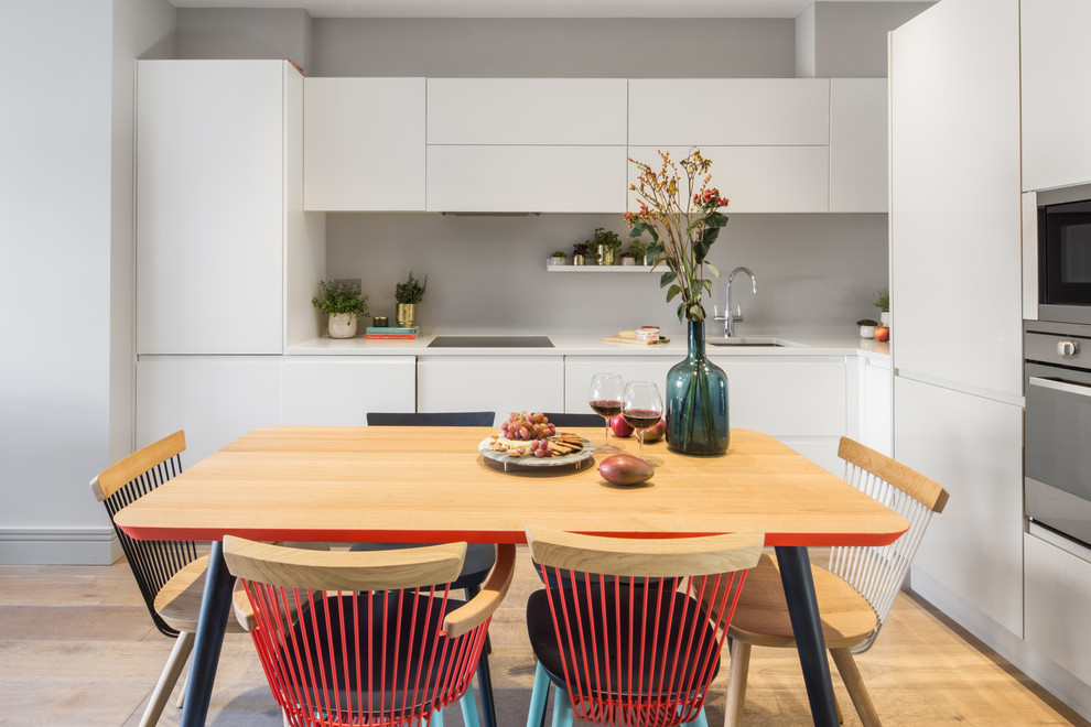 Photo of a scandinavian l-shaped eat-in kitchen in London with an undermount sink, flat-panel cabinets, white cabinets, grey splashback, stainless steel appliances, light hardwood floors and white benchtop.