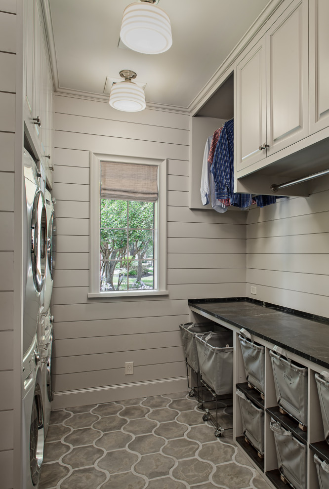 Photo of a mid-sized transitional galley dedicated laundry room in Houston with an utility sink, raised-panel cabinets, grey cabinets, soapstone benchtops, grey walls, concrete floors, a stacked washer and dryer, grey floor and black benchtop.