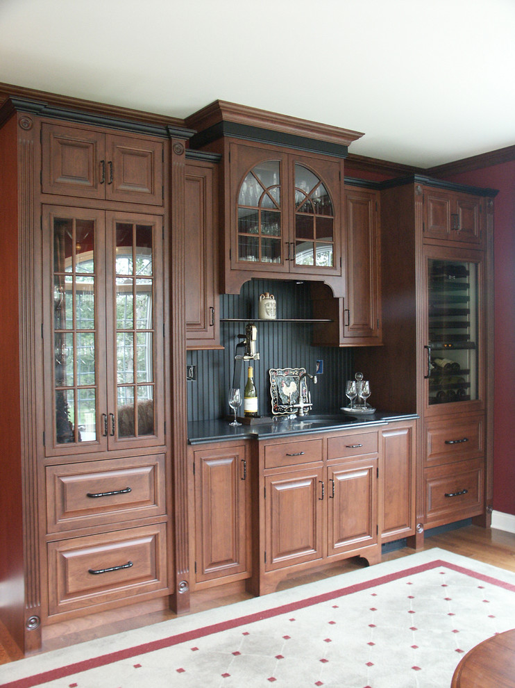 Photo of a mid-sized traditional wet bar in Philadelphia with an undermount sink, raised-panel cabinets, wood benchtops, black splashback, brown cabinets and medium hardwood floors.