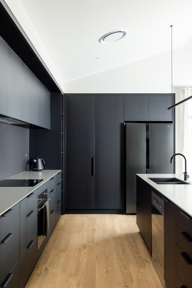 Photo of a small contemporary galley open plan kitchen in Christchurch with a submerged sink, black cabinets, composite countertops, black splashback, black appliances, laminate floors, an island, brown floors, grey worktops and a vaulted ceiling.