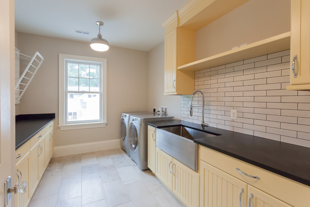 This is an example of a country galley dedicated laundry room in DC Metro with a farmhouse sink, beaded inset cabinets, yellow cabinets, beige walls, a side-by-side washer and dryer, beige floor and black benchtop.
