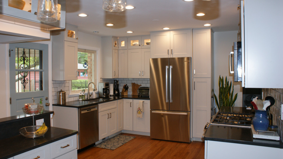 Photo of a mid-sized traditional u-shaped eat-in kitchen in Chicago with an undermount sink, flat-panel cabinets, grey cabinets, granite benchtops, white splashback, subway tile splashback, stainless steel appliances, medium hardwood floors, no island, brown floor and black benchtop.