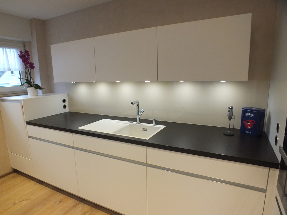 This is an example of a contemporary galley open plan kitchen in Dortmund with a drop-in sink, white cabinets, beige splashback, glass sheet splashback, light hardwood floors and with island.