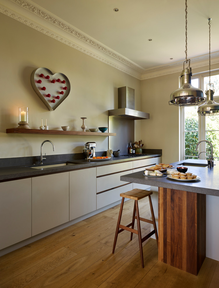 Large contemporary single-wall open plan kitchen in London with an integrated sink, flat-panel cabinets, white cabinets, limestone benchtops, light hardwood floors, with island, brown floor and black benchtop.