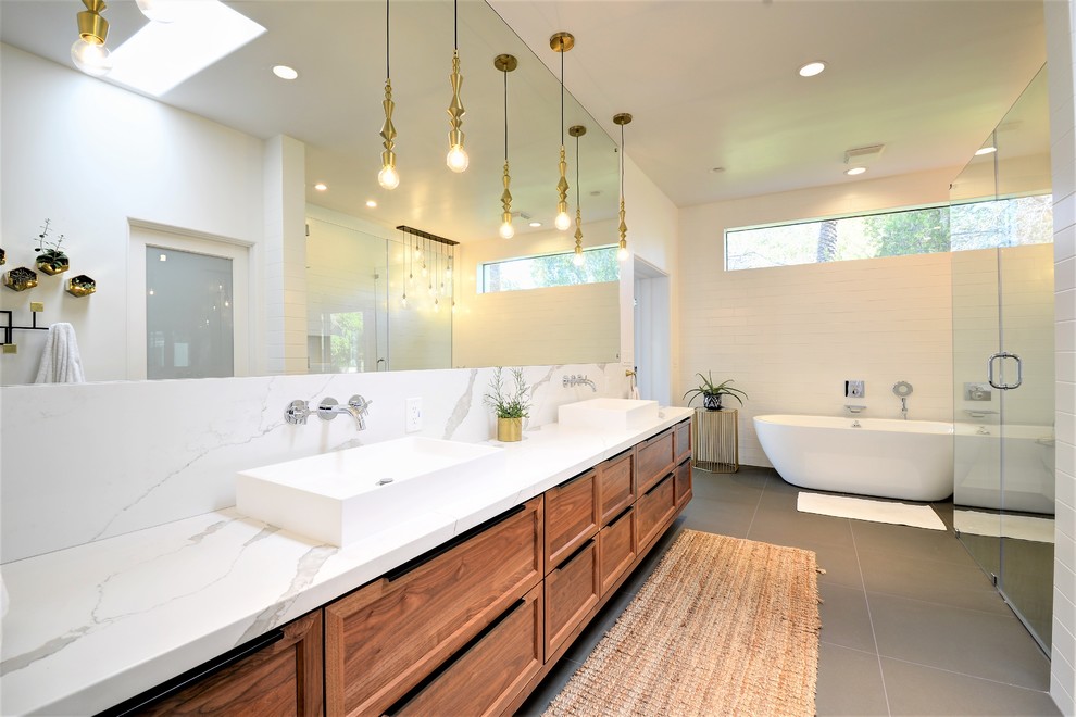 This is an example of a large contemporary master bathroom in Phoenix with shaker cabinets, medium wood cabinets, a freestanding tub, a curbless shower, white tile, white walls, porcelain floors, a vessel sink, granite benchtops, grey floor, a hinged shower door and white benchtops.
