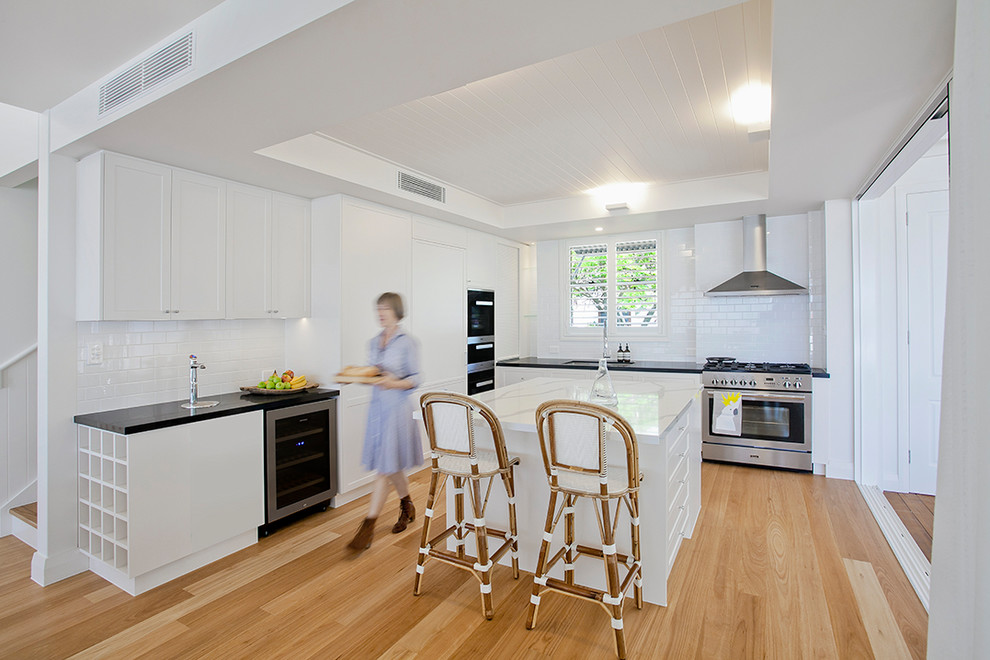 Large beach style l-shaped kitchen/diner in Sunshine Coast with an integrated sink, recessed-panel cabinets, white cabinets, granite worktops, white splashback, metro tiled splashback, integrated appliances, light hardwood flooring and an island.