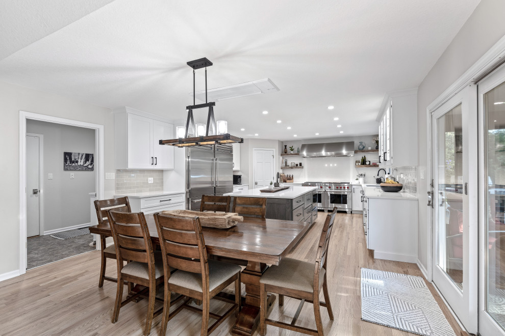 Example of a large transitional u-shaped light wood floor and multicolored floor eat-in kitchen design with a single-bowl sink, shaker cabinets, white cabinets, quartz countertops, white backsplash, quartz backsplash, stainless steel appliances, an island and white countertops