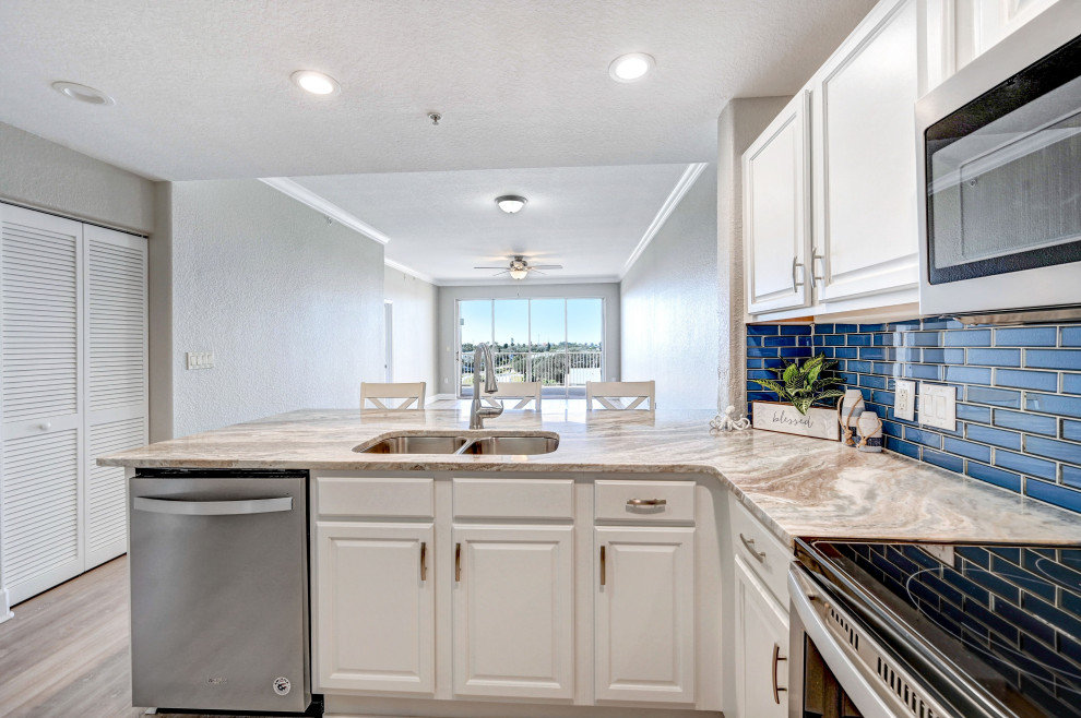 Mid-sized beach style u-shaped open plan kitchen in Orlando with a double-bowl sink, raised-panel cabinets, white cabinets, granite benchtops, blue splashback, glass tile splashback, stainless steel appliances, laminate floors, a peninsula, grey floor, multi-coloured benchtop and recessed.