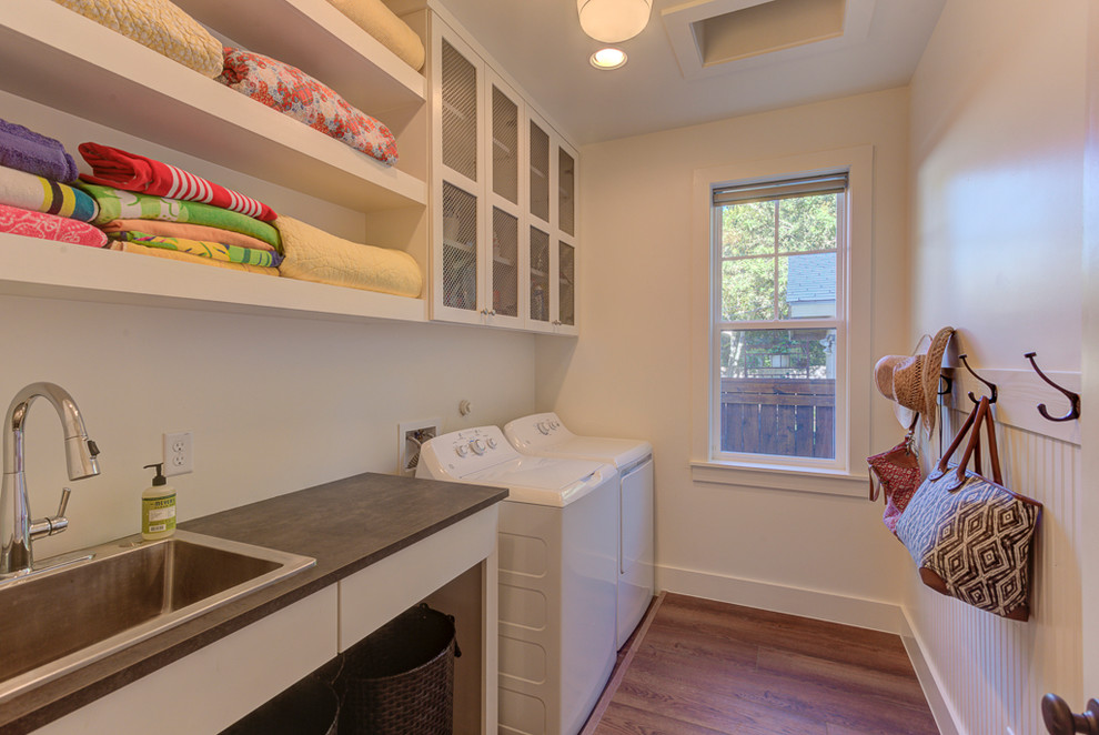 Photo of a small country galley dedicated laundry room in Austin with a drop-in sink, shaker cabinets, white cabinets, laminate benchtops, white walls, vinyl floors, a side-by-side washer and dryer, brown floor and black benchtop.