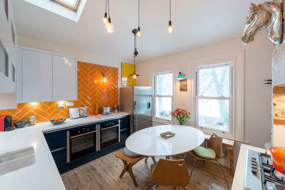 Inspiration for a mid-sized eclectic u-shaped eat-in kitchen in London with an undermount sink, flat-panel cabinets, black cabinets, orange splashback, subway tile splashback, stainless steel appliances, medium hardwood floors, no island, brown floor and white benchtop.