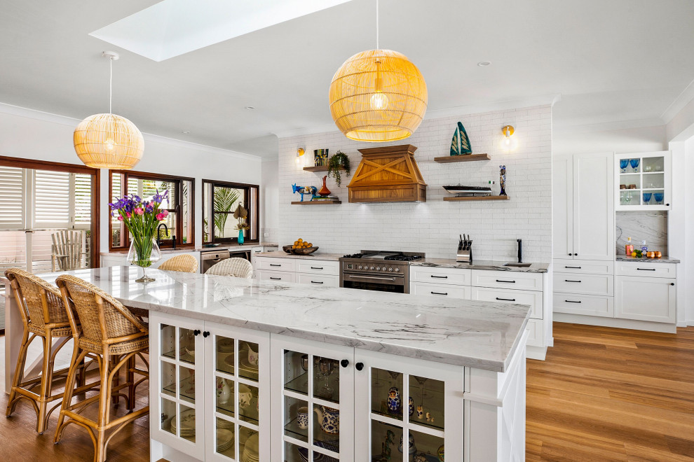 Photo of a tropical u-shaped kitchen in Sunshine Coast with an undermount sink, shaker cabinets, white cabinets, quartzite benchtops, white splashback, subway tile splashback, stainless steel appliances, medium hardwood floors, with island, brown floor and grey benchtop.