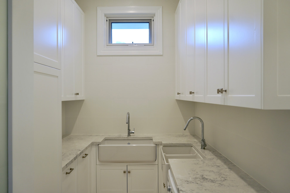 This is an example of a large modern u-shaped dedicated laundry room in Sydney with a farmhouse sink, shaker cabinets, white cabinets, quartz benchtops, white splashback, porcelain splashback, white walls, marble floors, a side-by-side washer and dryer, white floor and white benchtop.