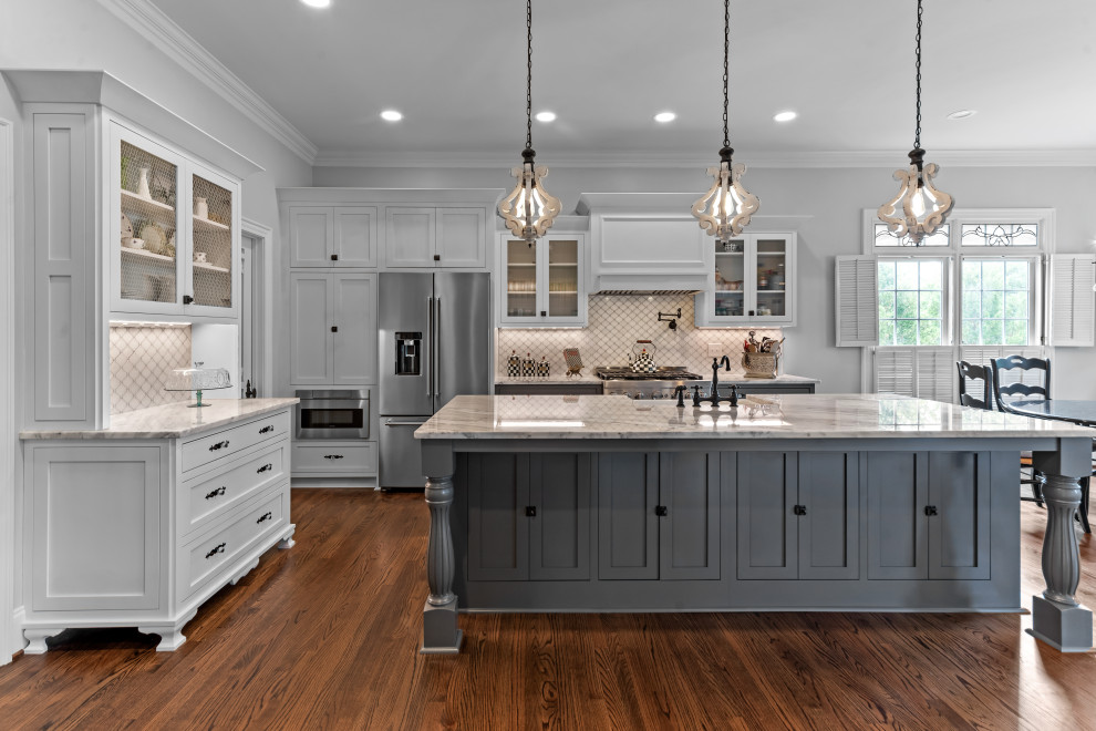 Photo of a country l-shaped kitchen in Raleigh with a farmhouse sink, shaker cabinets, white cabinets, white splashback, stainless steel appliances, dark hardwood floors, with island, brown floor and grey benchtop.