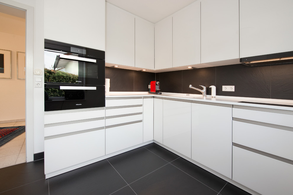 Small contemporary l-shaped eat-in kitchen in Stuttgart with an integrated sink, glass-front cabinets, white cabinets, solid surface benchtops, black splashback, black appliances, slate floors, no island, stone slab splashback and black floor.