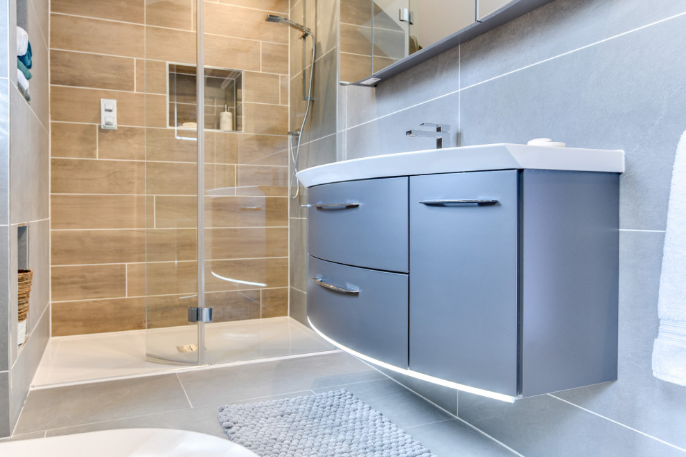 Inspiration for a large modern grey and white ensuite bathroom in Sussex with flat-panel cabinets, grey cabinets, a freestanding bath, a walk-in shower, a one-piece toilet, grey tiles, ceramic tiles, grey walls, ceramic flooring, a built-in sink, glass worktops, grey floors, an open shower, white worktops, feature lighting, a single sink and a floating vanity unit.