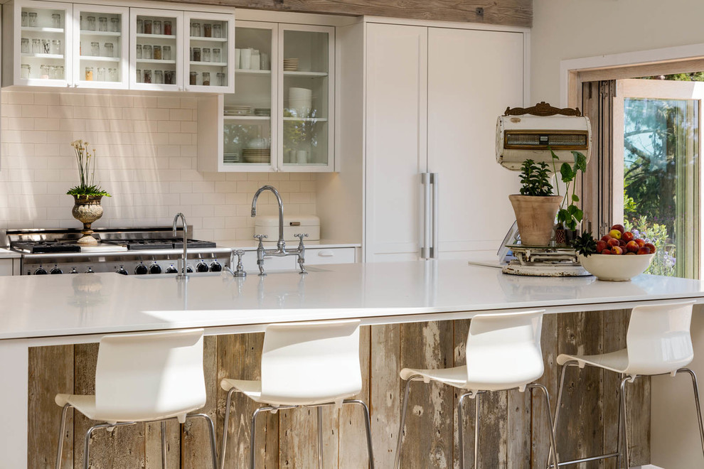 This is an example of a country kitchen in Los Angeles with a farmhouse sink, glass-front cabinets, white cabinets, white splashback, subway tile splashback, stainless steel appliances, with island and white benchtop.