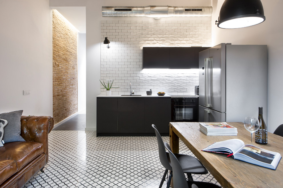 This is an example of a mid-sized industrial single-wall open plan kitchen in Other with flat-panel cabinets, black cabinets, white splashback, subway tile splashback, black appliances, ceramic floors, no island and multi-coloured floor.
