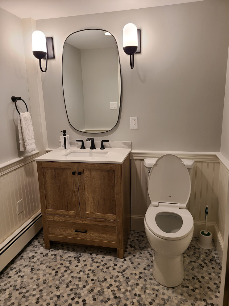 Inspiration for a medium sized urban bathroom in Boston with recessed-panel cabinets, medium wood cabinets, a two-piece toilet, grey walls, pebble tile flooring, a submerged sink, marble worktops, multi-coloured floors, white worktops, a single sink, a freestanding vanity unit and wainscoting.