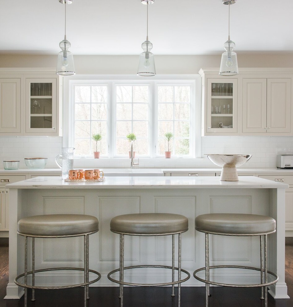This is an example of a beach style kitchen in New York with a farmhouse sink, beige cabinets, white splashback, subway tile splashback, dark hardwood floors, with island and beaded inset cabinets.