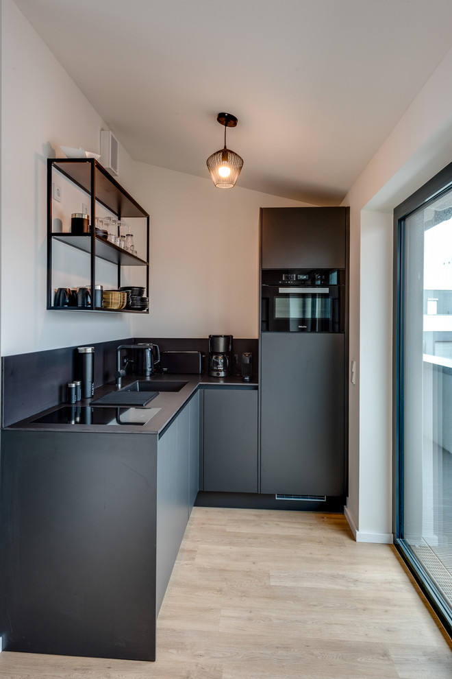 Inspiration for a small contemporary l-shaped open plan kitchen in Hamburg with a drop-in sink, flat-panel cabinets, grey cabinets, laminate benchtops, grey splashback, mirror splashback, black appliances, light hardwood floors, no island, beige floor and grey benchtop.