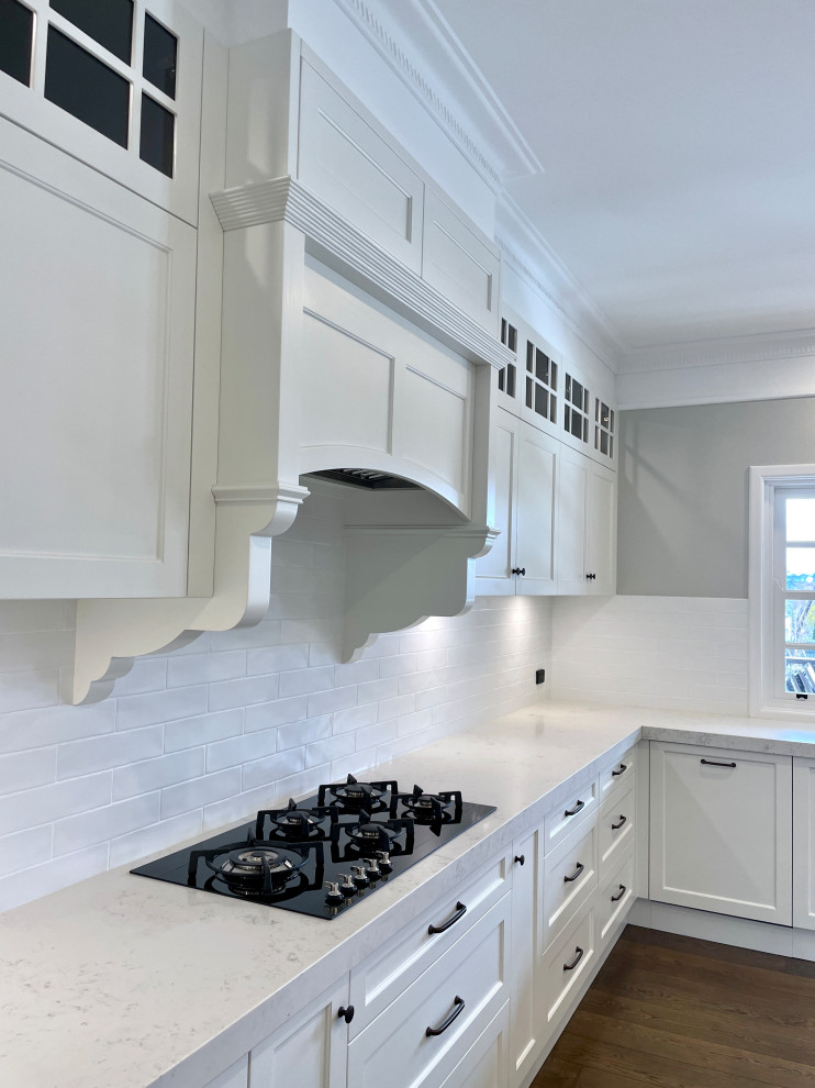 Bild på ett mycket stort vintage vit vitt kök, med en undermonterad diskho, luckor med infälld panel, vita skåp, bänkskiva i kvarts, vitt stänkskydd, stänkskydd i tunnelbanekakel, svarta vitvaror, mellanmörkt trägolv, en köksö och brunt golv