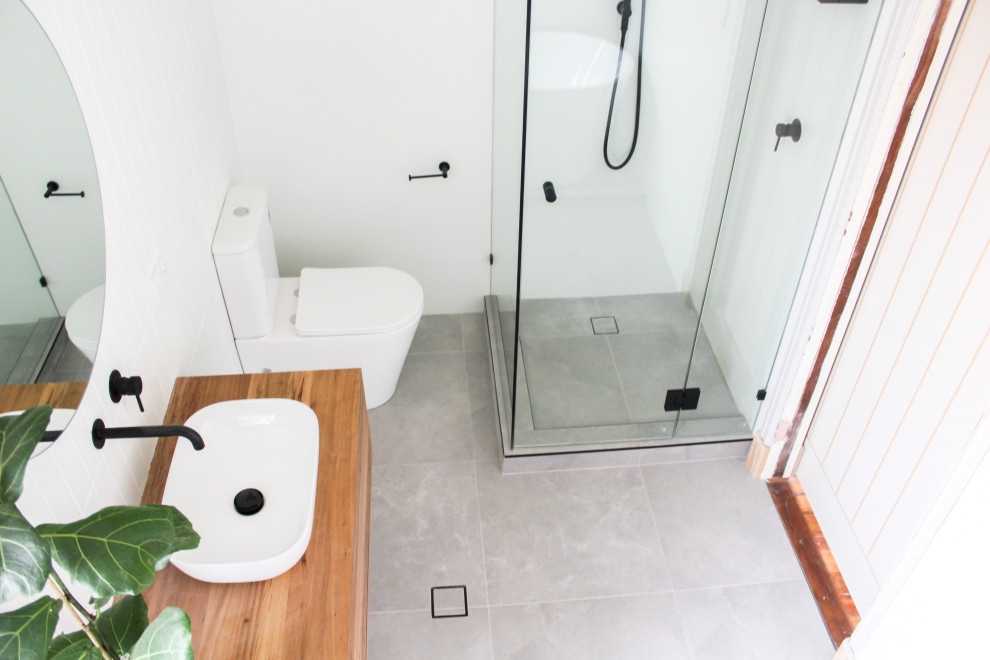 This is an example of a large modern master bathroom in Perth with furniture-like cabinets, medium wood cabinets, a freestanding tub, a corner shower, a one-piece toilet, white tile, subway tile, white walls, porcelain floors, a vessel sink, wood benchtops, grey floor, a hinged shower door, multi-coloured benchtops, a single vanity and a floating vanity.