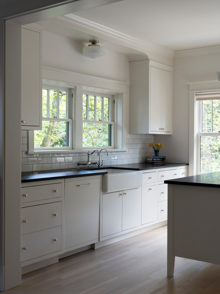 Example of a transitional light wood floor kitchen design in DC Metro with a farmhouse sink, white cabinets, granite countertops, white backsplash, ceramic backsplash, paneled appliances, an island and black countertops