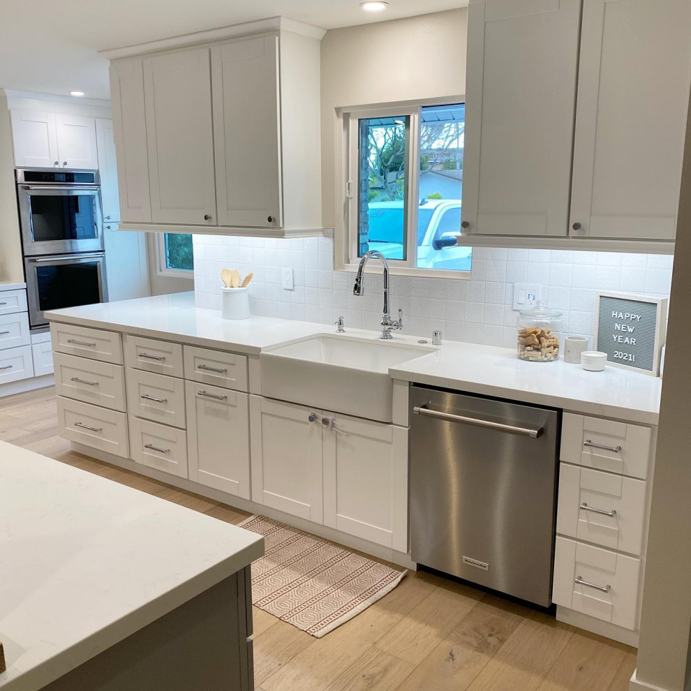 Large contemporary l-shaped open plan kitchen in Orange County with a farmhouse sink, shaker cabinets, white cabinets, quartz benchtops, white splashback, ceramic splashback, stainless steel appliances, light hardwood floors, with island, brown floor and white benchtop.