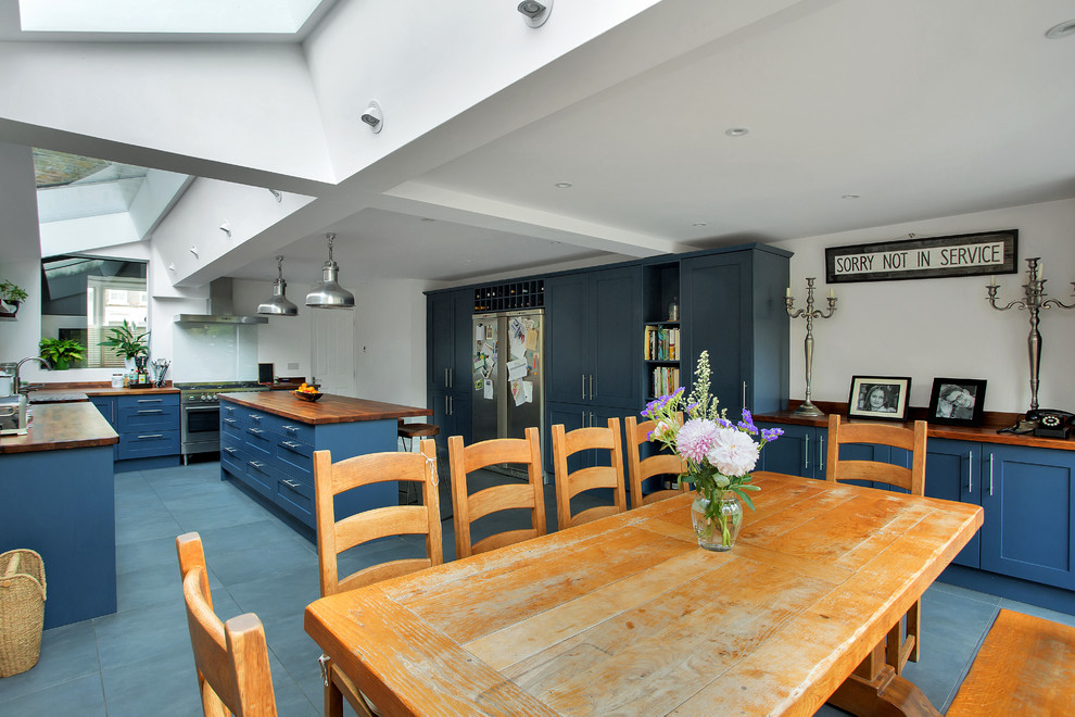 Large industrial eat-in kitchen in London with a farmhouse sink, shaker cabinets, blue cabinets, wood benchtops, white splashback, glass sheet splashback, stainless steel appliances, ceramic floors, with island, grey floor and brown benchtop.
