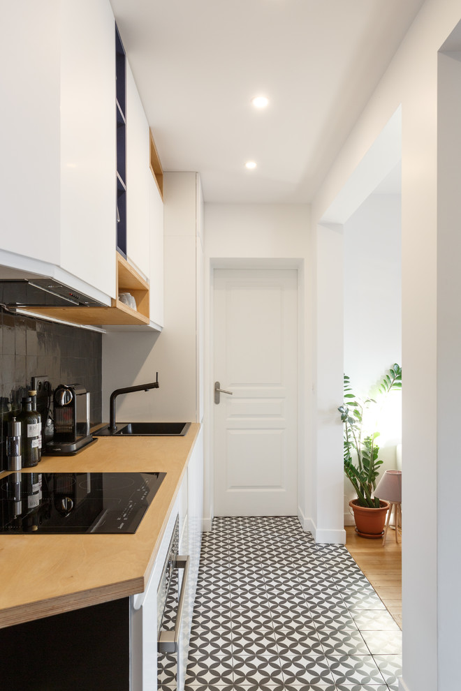 This is an example of a small modern single-wall open plan kitchen in Paris with a drop-in sink, flat-panel cabinets, white cabinets, wood benchtops, black splashback, terra-cotta splashback, stainless steel appliances, cement tiles, no island, multi-coloured floor and beige benchtop.