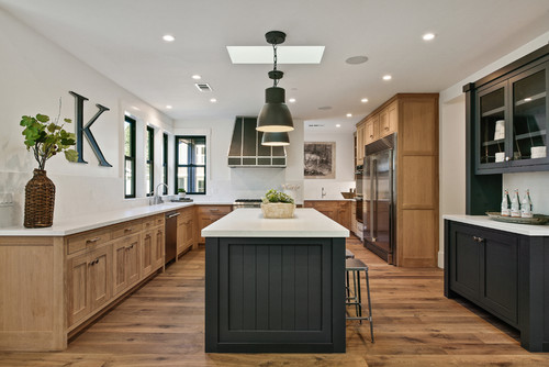 Modern open-concept kitchen with two-tone kitchen cabinet design, featuring natural wood perimeter cabinets, a dark island, and stainless steel appliances on wide plank wood flooring.