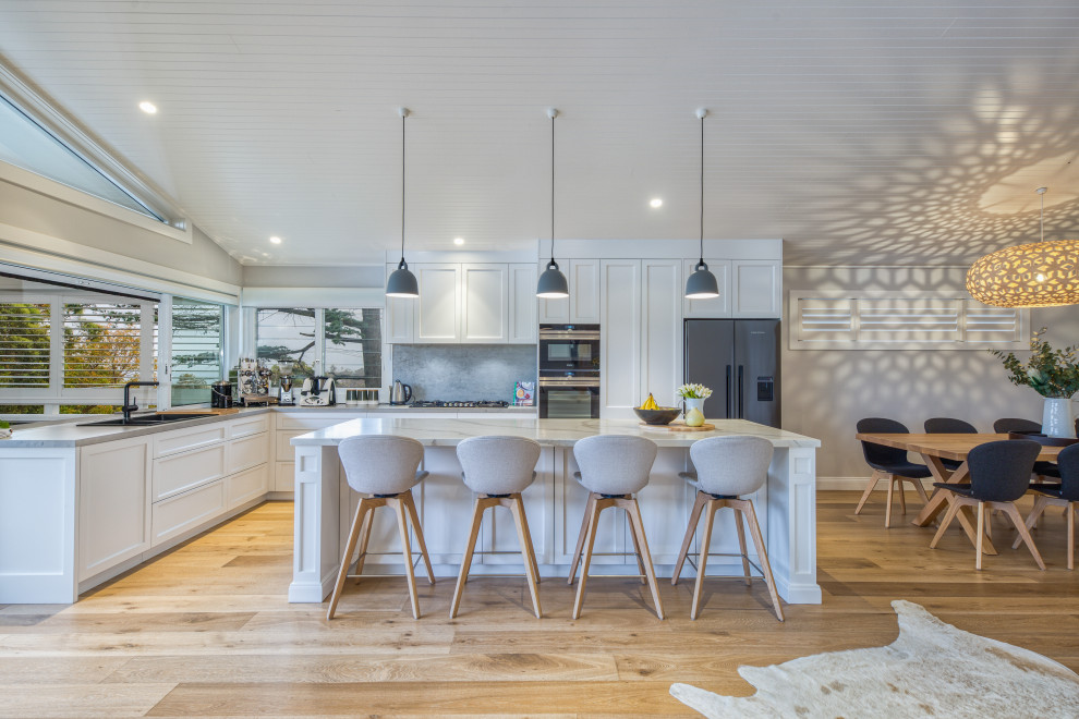 Design ideas for a large beach style l-shaped kitchen in Sydney with a drop-in sink, shaker cabinets, white cabinets, grey splashback, stainless steel appliances, light hardwood floors, with island, beige floor and white benchtop.