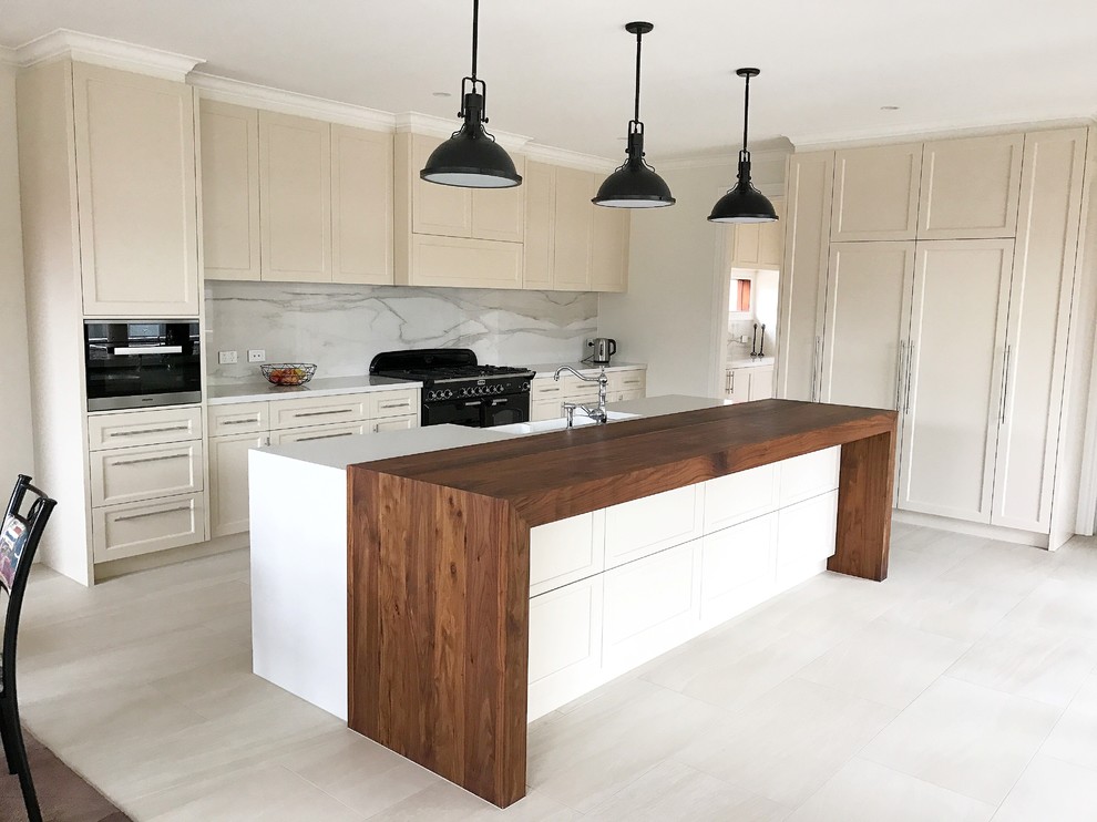 Photo of a mid-sized traditional galley open plan kitchen in Melbourne with a farmhouse sink, shaker cabinets, beige cabinets, white splashback, porcelain splashback, black appliances, porcelain floors, with island and wood benchtops.