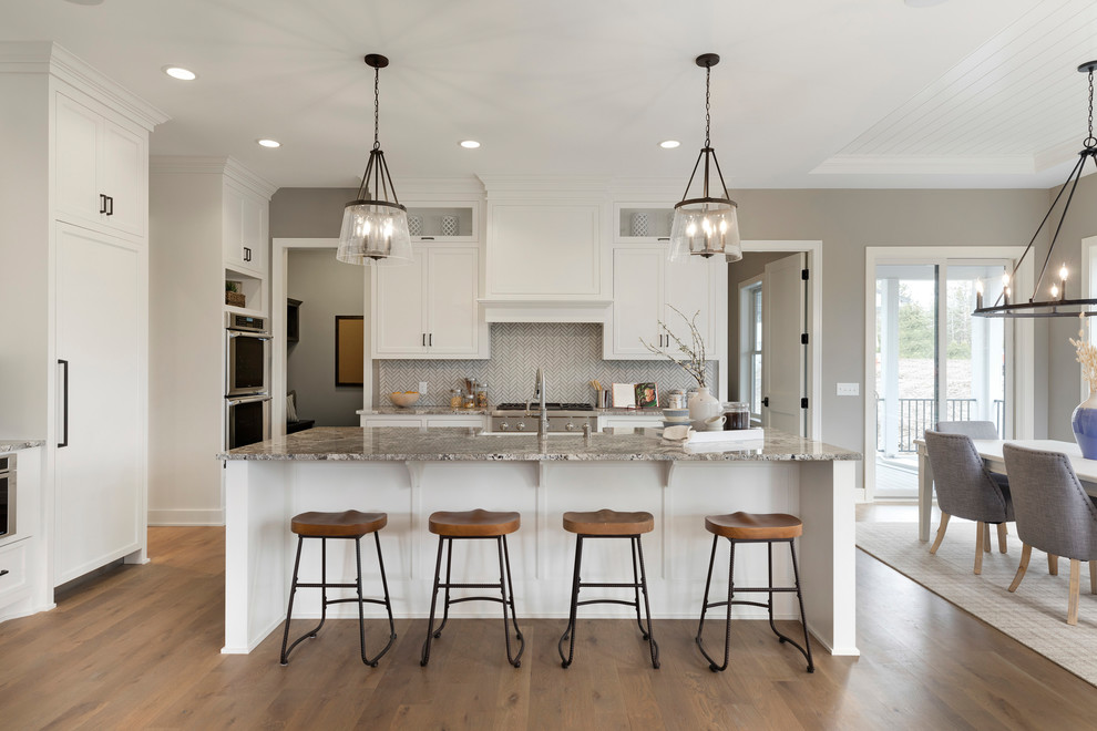 Inspiration for a large transitional l-shaped open plan kitchen in Minneapolis with a farmhouse sink, shaker cabinets, white cabinets, granite benchtops, grey splashback, mosaic tile splashback, stainless steel appliances, medium hardwood floors, with island, multi-coloured benchtop and brown floor.