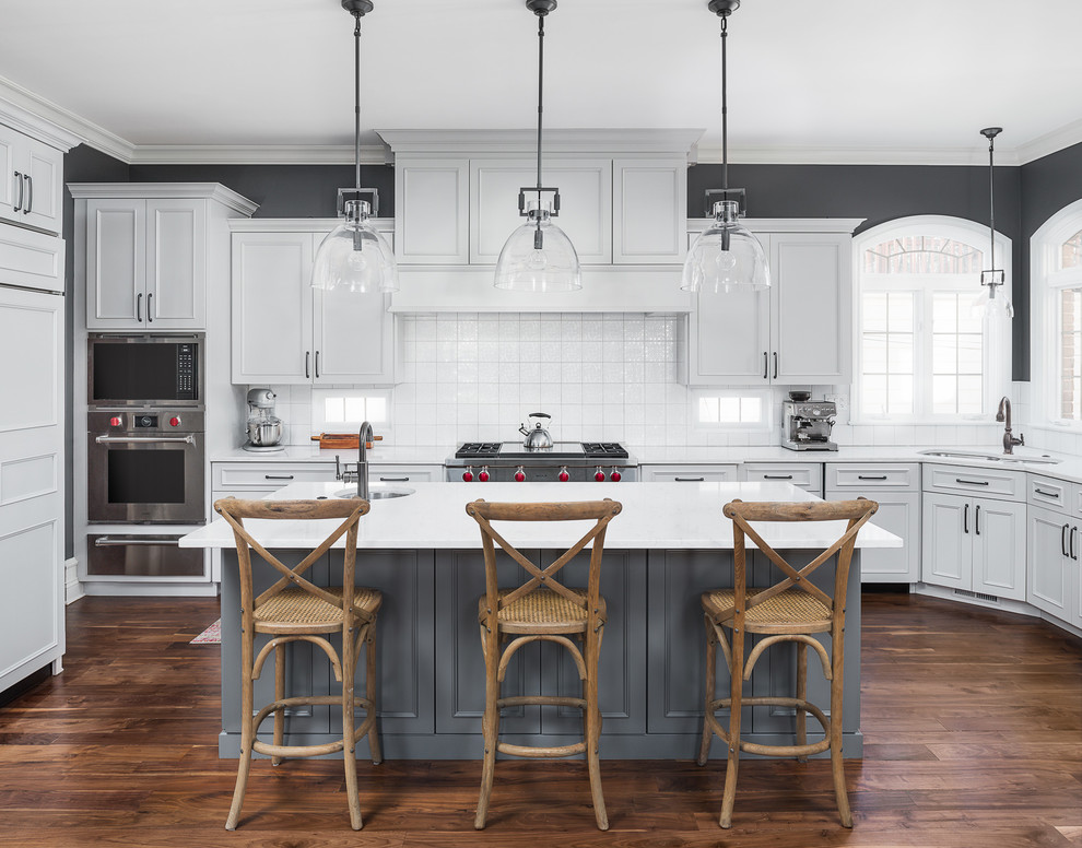 Inspiration for a large transitional l-shaped medium tone wood floor and brown floor eat-in kitchen remodel in Chicago with an undermount sink, white cabinets, quartz countertops, white backsplash, ceramic backsplash, stainless steel appliances, an island and white countertops