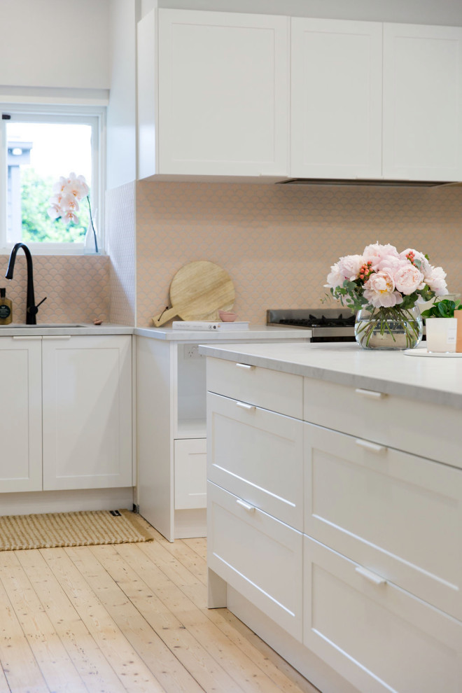 This is an example of a small scandinavian l-shaped eat-in kitchen in Melbourne with an undermount sink, recessed-panel cabinets, white cabinets, quartz benchtops, pink splashback, mosaic tile splashback, black appliances, medium hardwood floors, with island and white benchtop.