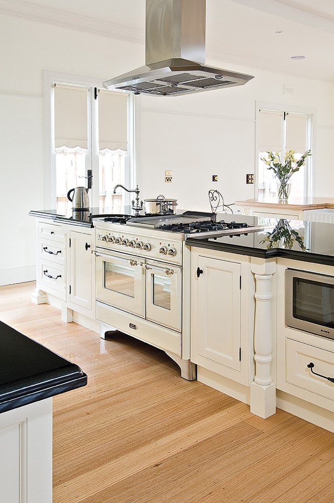This is an example of a large traditional galley open plan kitchen in Sydney with a farmhouse sink, recessed-panel cabinets, white cabinets, white appliances, light hardwood floors and multiple islands.