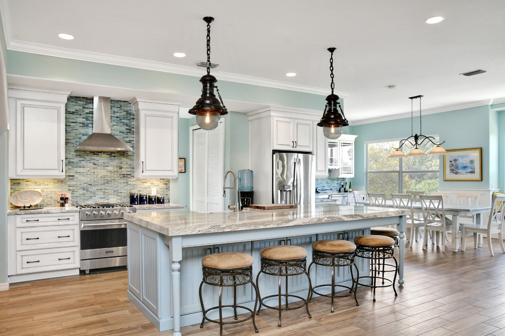 Design ideas for a large beach style l-shaped eat-in kitchen in Tampa with a farmhouse sink, raised-panel cabinets, white cabinets, quartz benchtops, blue splashback, glass tile splashback, stainless steel appliances, porcelain floors and with island.