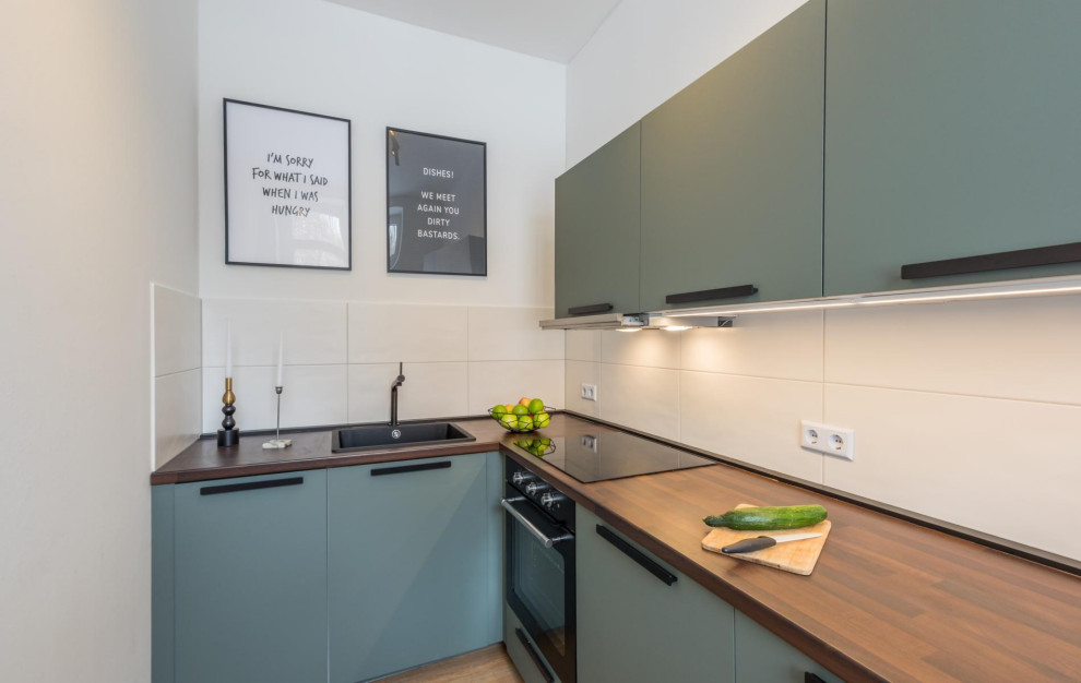 Small modern l-shaped open plan kitchen in Dresden with a single-bowl sink, beaded inset cabinets, green cabinets, wood benchtops, white splashback, ceramic splashback, panelled appliances, light hardwood floors, no island, brown floor, brown benchtop and wallpaper.