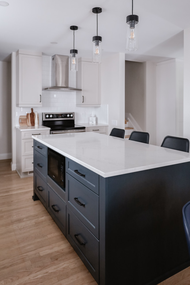 Example of a mid-sized transitional l-shaped light wood floor and brown floor eat-in kitchen design with an undermount sink, shaker cabinets, beige cabinets, quartz countertops, white backsplash, subway tile backsplash, stainless steel appliances, an island and white countertops